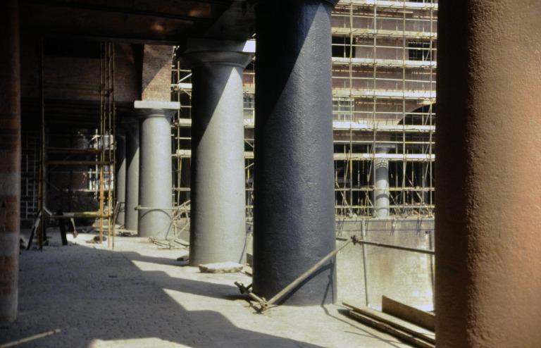 Photograph of renovation of Albert Dock, Liverpool, Block D, later Merseyside Maritime Museum, colonnade, quayside on south side, looking east towards scaffolded Block E.  View of cast-iron columns showing three stages of paint finishes. card